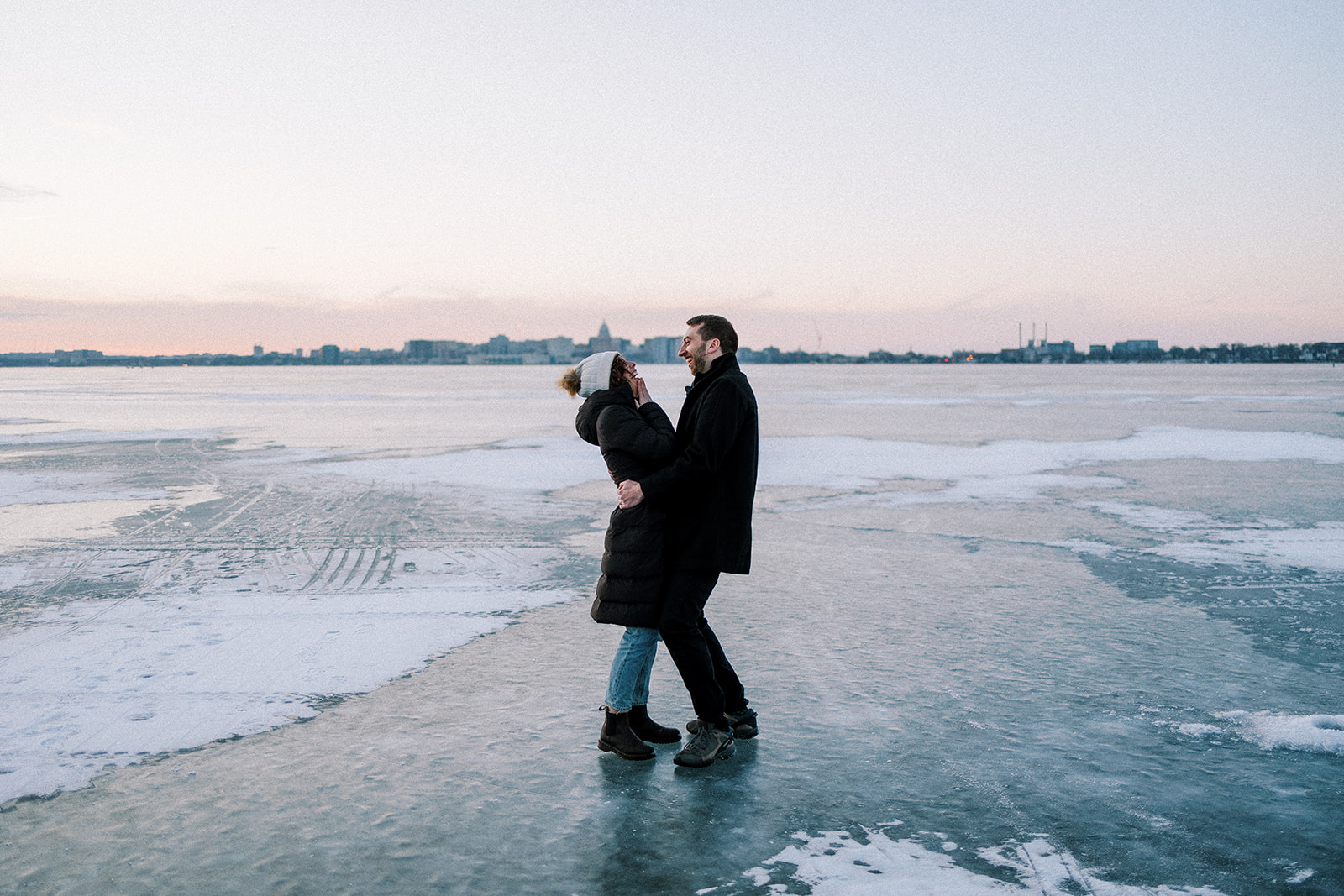 couple stands on frozen lake in madison wisconsin and laughs as they hold each other