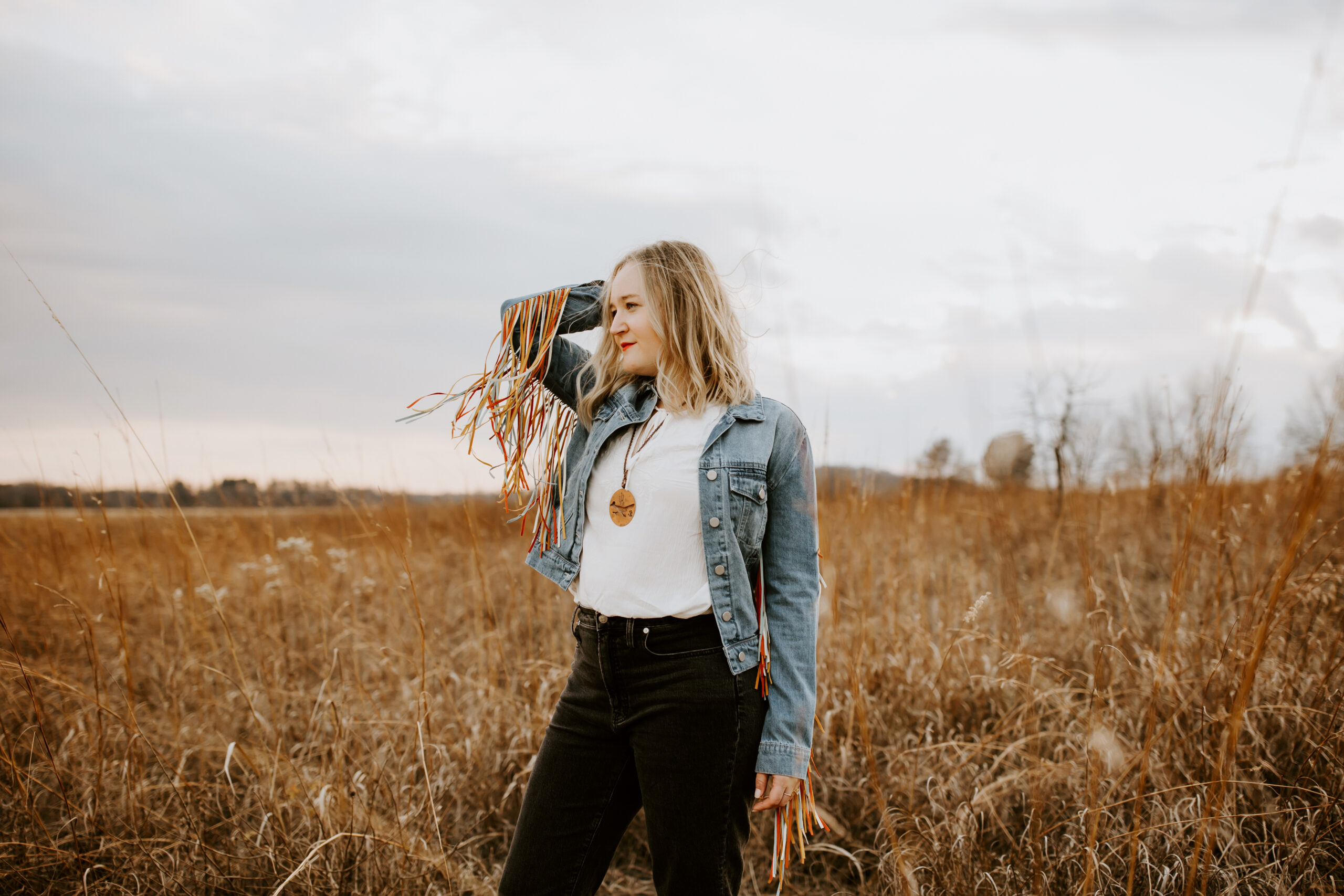 Anya Kubilus stands in field with fringe jacket and wind blowing in her hair